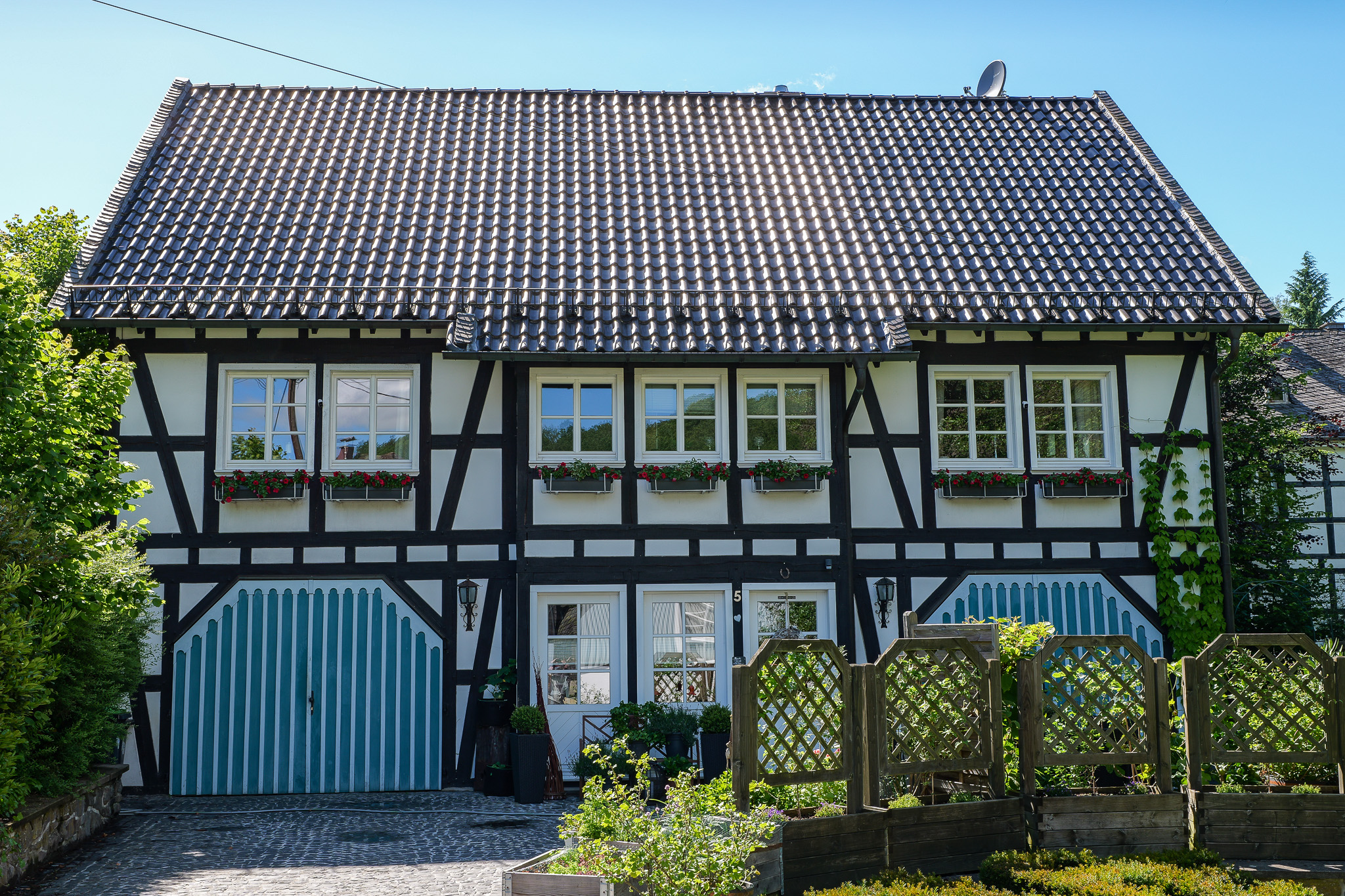 Traditionelles Fachwerkhaus in Holpe mit schwarzen Balken auf weißem Untergrund, roten Blumenkästen an den Fenstern, blauer Holztür und gepflegtem Vorgarten mit Rankgitter und Bepflanzung.