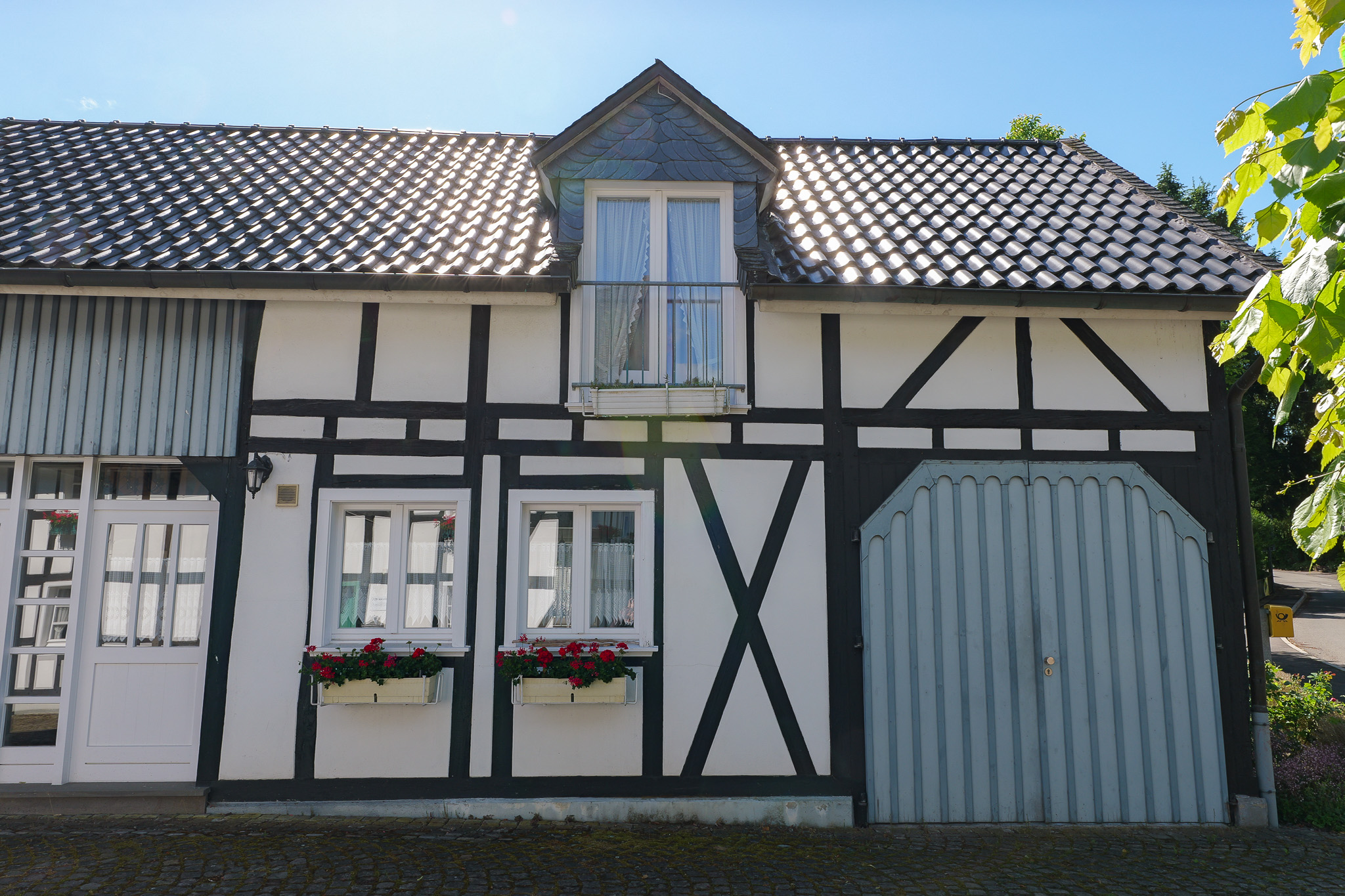 Fachwerkhaus in Holpe mit schwarzen Balken auf weißem Untergrund, großem grau-blauem Scheunentor, roten Blumenkästen an den Fenstern, einem Dachgauben-Balkon im Obergeschoss und Weinranken an der Hauswand.